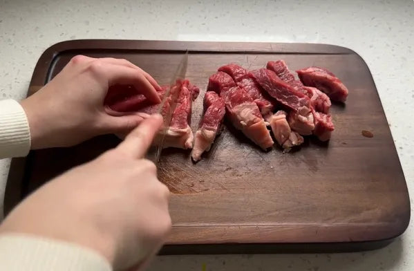 Hands cutting raw red meat into strips on a dark wooden cutting board